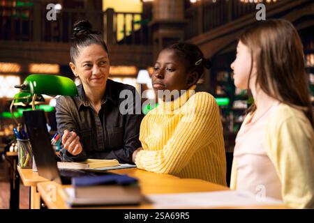 Preteen scholars collaborating with their tutor in a learning environment, working on school projects and developing critical classroom skills. Woman mentoring her students in the library. Stock Photo