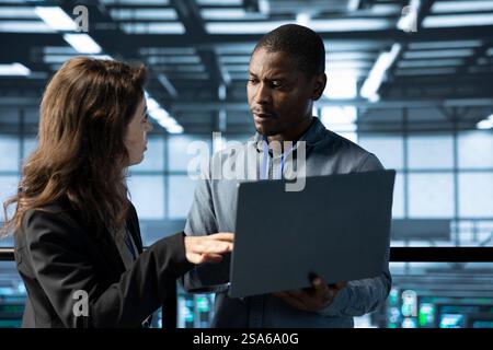 System administrators in data center using laptop, analyzing metrics, optimizing storage volumes. Server farm IT experts using notebook, talking, ensuring seamless operations Stock Photo