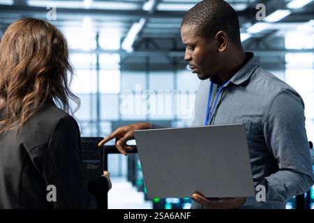 Colleagues in data center workspace use laptop to calibrate energy usage. Server room IT specialist using notebook to inspect UPS configurations, doing maintenance on equipment Stock Photo