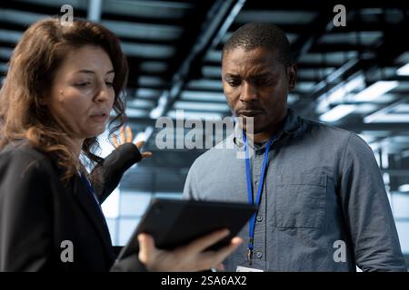 Team of engineers in data center talking, using tablet to do network virtualization. Coworkers doing brainstorming in server farm, updating storage arrays with device, verifying settings Stock Photo