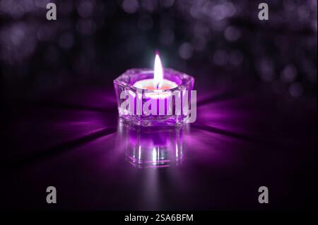 lit purple candle in a glass container reflecting on a table and with bokeh in the background Stock Photo