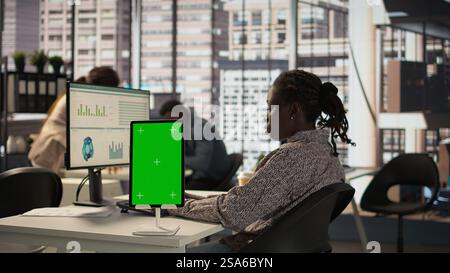 Female manager seated at workstation with green screen on devices, working on solving administrative tasks for enterprise growth. Developing performance metrics to support firm goals. Camera B. Stock Photo