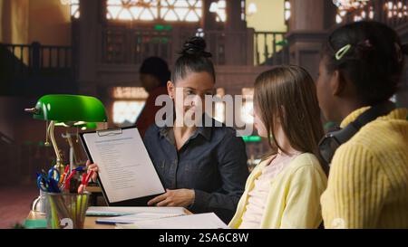 Female mentor providing instruction to diverse pupils during private lessons at the library, assisting with homework and school projects in the study area. Tutoring session for guidance. Camera A. Stock Photo
