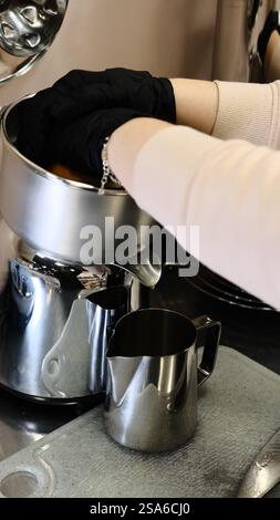 A barista in black disposable gloves squeezes fresh oranges on a professional juicer into a metal pitcher in a modern kitchen preparing a refreshing Stock Photo
