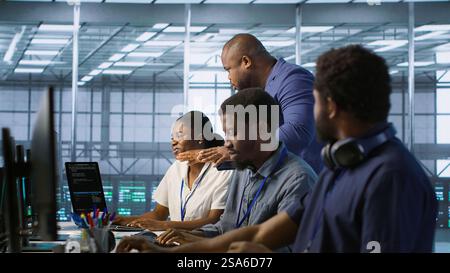 Data center team leader supervising engineers implementing AI predictive maintenance systems. Server farm boss overseeing workers using artificial intelligence to identify hardware failures, camera A Stock Photo