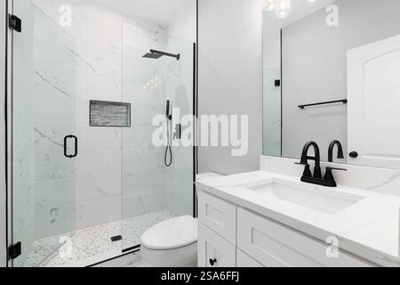 A bathroom with a white cabinet and marble countertop, black faucet and showerhead, and a marble tiled shower with a stone tiled floor. Stock Photo