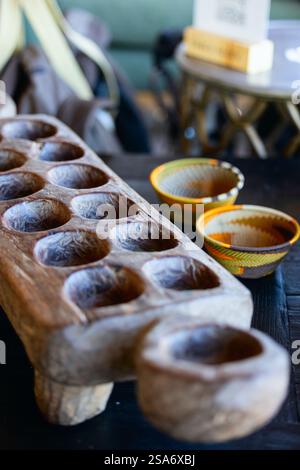 A close-up of a traditional wooden Mancala board with carved pits, accompanied by colorful woven bowls in the background. Represents African cultural Stock Photo