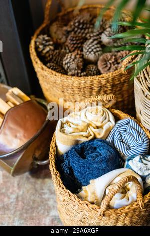 Natural woven baskets containing folded scarves in blue and cream colors, alongside decorative pine cones and indoor plant. Stock Photo