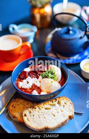 Mediterranean shakshuka served in blue bowl with poached eggs, feta cheese, microgreens, and chorizo, accompanied by sliced sourdough. Stock Photo