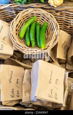 Four fresh green zucchini displayed in rustic wicker basket, surrounded by brown paper bags with handwritten market labels. Stock Photo