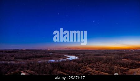 Panorama of constellation Orion and the winter stars setting into the evening twilight sky oner Bow River valley in southern Alberta, Canada. Orion is left of centre, with the bright star Sirius in Canis Major at far left. At right is Aldebaran amid the Hyades star cluster, and the smaller Pleiades star cluster to the right, all in Taurus. At lower right is Jupiter. The Belt stars of Orion point left (south) to Sirius and right (north) to Aldebaran.   The scene is lit by the twilight but also by light from the waxing gibbous Moon off frame to the left. Photographed on 19 April 2024. Stock Photo