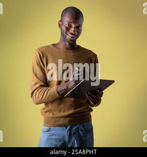 Smiling African man interacting with a digital tablet using a stylus, standing against a yellow background. Perfect for technology, creativity, and pr Stock Photo