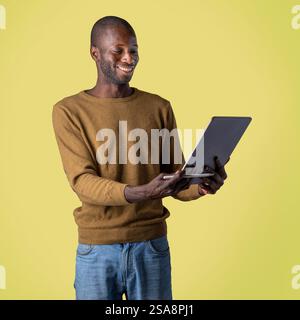 Cheerful Black man holding a digital tablet with a blank screen, wearing a brown sweater against a vibrant yellow background, technology and presentat Stock Photo
