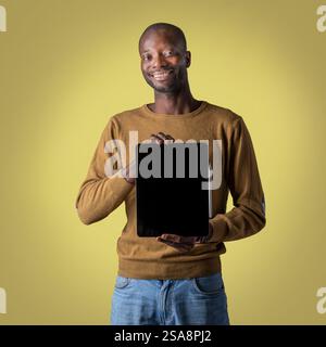 Smiling African man in casual outfit presenting a blank tablet with a black screen, standing against a yellow background, suitable for advertising or Stock Photo