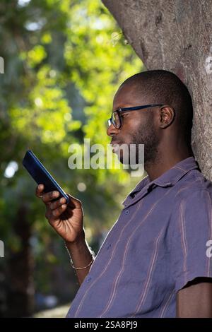 Young African man using his smartphone to send a voice message in hands-free mode while leaning on a tree outdoors. Focus on digital communication and Stock Photo