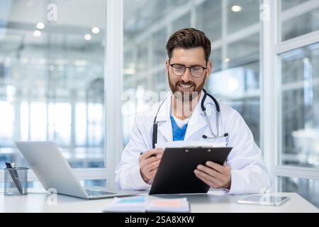 Successful experienced doctor records patient data using flipchart, man in white medical coat at workplace inside clinic, prepares patient treatment report. Stock Photo