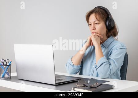 Serious freelance professional woman wearing big headphones listening and watching video, online conference, video meeting, webinar or studying at the Stock Photo