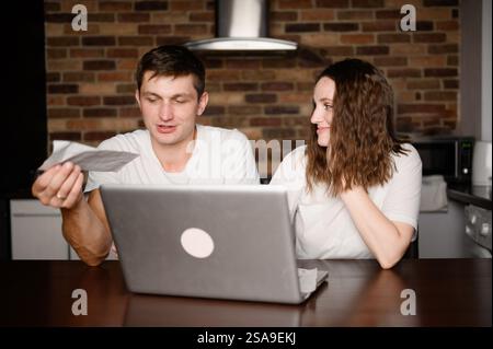 Young wife and husband are sitting at home with laptop, papers, looking at documents, checks, analyzing expenses together. Analysis of money spending Stock Photo