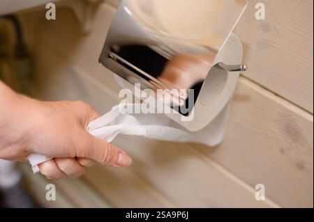 Woman tears piece of toilet paper from roll hanging on holder on wall, close-up. Reasonable consumption. Concept of health problems, intestines, perso Stock Photo