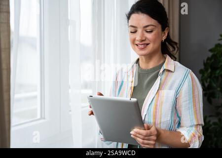 Smart caucasian woman in business casual wear holding digital tablet standing near windows at home. Young freelance independent expert female worker u Stock Photo