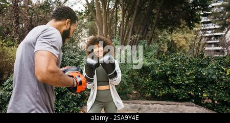 A cheerful woman wearing boxing gloves practices boxing with a trainer in a park. Surrounded by greenery, they enjoy fitness and activity in a urban p Stock Photo