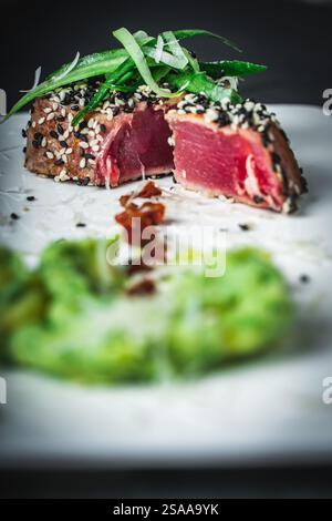 Close-up of a delicious tuna steak in a sesame crust. Medium-rare tuna slices with green puree and sun-dried tomatoes on a white plate on a dark backg Stock Photo