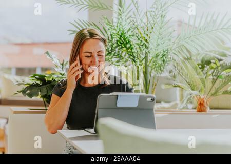 A woman is talking on her cell phone while sitting at a table with a tablet. She is smiling and she is enjoying her conversation. The table is surroun Stock Photo