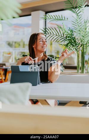 A woman is sitting at a table with a tablet and a pen working and pointing to the right. Stock Photo