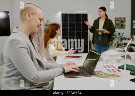 Group of professionals discussing renewable energy solutions using laptops and solar panel model for presentations in modern office setting Stock Photo