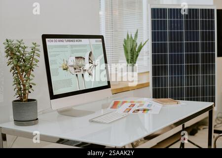 Displaying computer screen with detailed wind turbine diagram on workstation. Desk showing potted plants, solar panel, and papers with charts Stock Photo