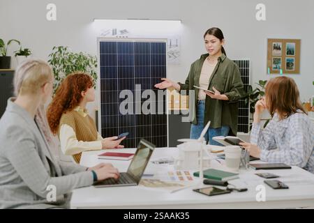 Team of professionals discussing renewable energy in office with solar panel on display, using various gadgets and devices and collaborating on project Stock Photo