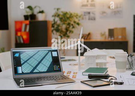 Workspace setting featuring laptop displaying solar panel design and miniature wind turbine model While office setup indicates a focus on sustainable and renewable energy projects Stock Photo