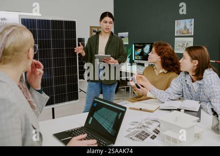 Woman presenting renewable energy project to diverse team using a tablet and solar panel display. Group of colleagues listening attentively and discussing technical details Stock Photo
