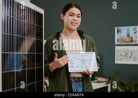 Woman holding tablet while standing next to solar panel and presentation board She is explaining renewable energy solutions with graphical data display Stock Photo