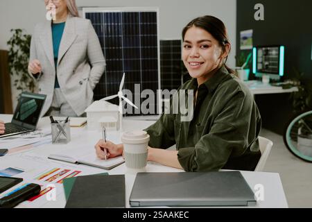 Hispanic woman smiling while working on sustainable project demonstrating renewable energy models in modern office environment engaged in team collaboration Stock Photo