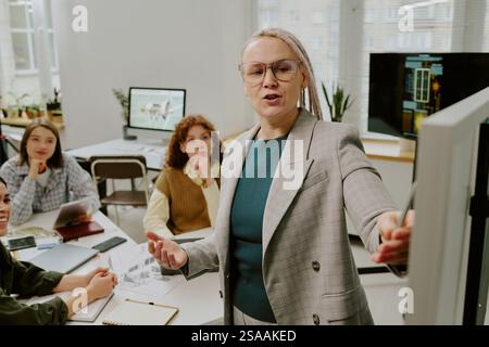 Portrait of middle-aged woman in grey suit conducting business presentation in office setting. Diverse team of colleagues engaging and listening attentively Stock Photo