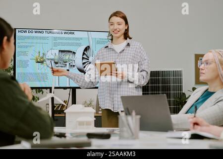 Young woman explaining wind turbine functionality to colleagues in modern office. Team members engaged in discussion while viewing presentation on large screen Stock Photo