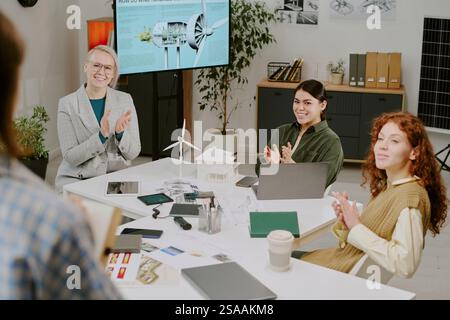 Group of professionals applauding colleague during presentation in modern office setting featuring charts, laptop, and model wind turbine Stock Photo