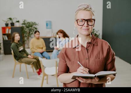 Portrait of woman holding notebook while standing in classroom with students. Classroom environment includes educational tools and background elements suggesting active learning Stock Photo