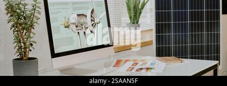 Modern desktop computer displaying renewable energy details on screen. Desk organized with potted plant, booklets, and solar panel in background Stock Photo