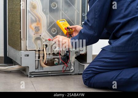 A photo of a young man using an electronic multimeter while repairing a dishwasher in a house Stock Photo