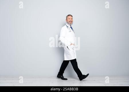 Smiling male doctor walking confidently in white coat with hands in pockets, gray background emphasizing calm professionalism Stock Photo
