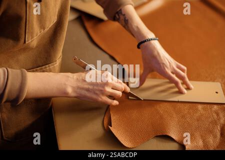 Close-up of hands skillfully cutting leather with a blade, showing detailed craftsmanship. Table covered with pieces of leather and tools for crafting Stock Photo
