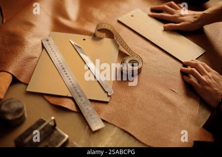 Artisan hands working on leather with precision tools, creating customized leather goods on a wooden table. Measuring tape, ruler, and cutting blade are arranged nearby Stock Photo
