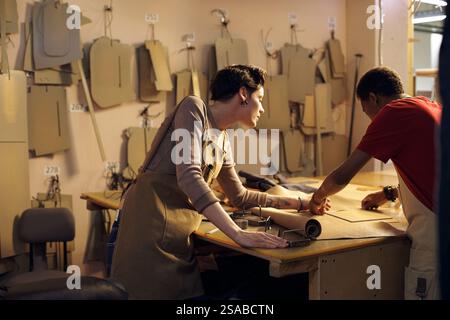 Two people crafting intricate patterns in leather workshop, surrounded by various tools and materials hanging on wall. Focus is on collaboration and detailed work Stock Photo