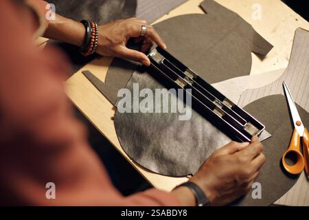 Hands working with precision while cutting piece of leather on workbench using specialized cutting tool. Surroundings include other materials and tools important for leatherworking Stock Photo