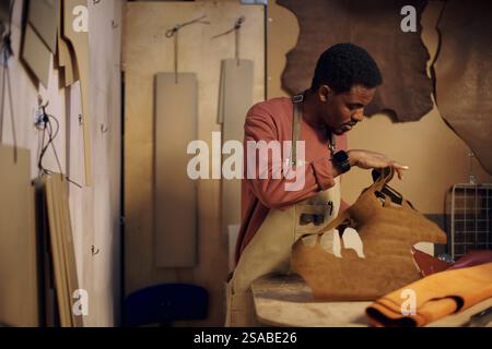 Skilled artisan working in workshop while carefully crafting leather goods. Craftsman is seen engrossed in intricate work, surrounded by various tools and leather pieces Stock Photo