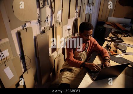 Person working on laptop in workshop surrounded by various materials and tools creating detailed models for design project, using digital assistance in organized workspace focused on task Stock Photo