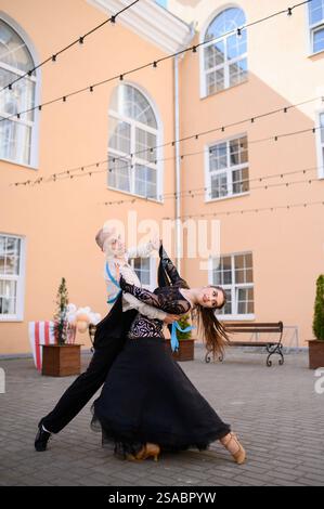 Beautiful couple of two professional ballroom dancers dancing outside. Sport life, music, classical dance concept. Young woman in black dress and man in white shirt dancing tango at performance Stock Photo