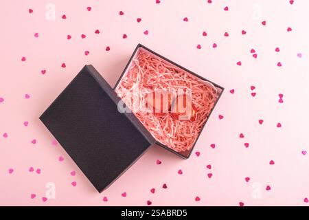 Open black gift box with red heart-shaped chocolates and pink shredded paper on a pastel pink background. Valentines day concept. Top view Stock Photo